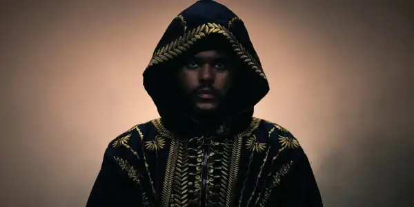 The Weeknd w czarnej, haftowanej złotem kurtce z kapturem, patrzący prosto w obiektyw.