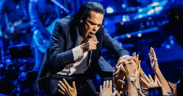 Nick Cave w garniturze śpiewa do mikrofonu, otoczony przez wyciągnięte ręce fanów.