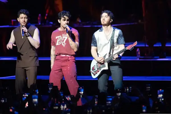 Trzech członków zespołu Jonas Brothers na scenie, jeden śpiewa, drugi gra na gitarze.