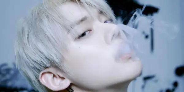 Młody mężczyzna z jasnymi włosami, jeden z członków popularnych k-pop zespołów męskich, wydmuchuje dym.