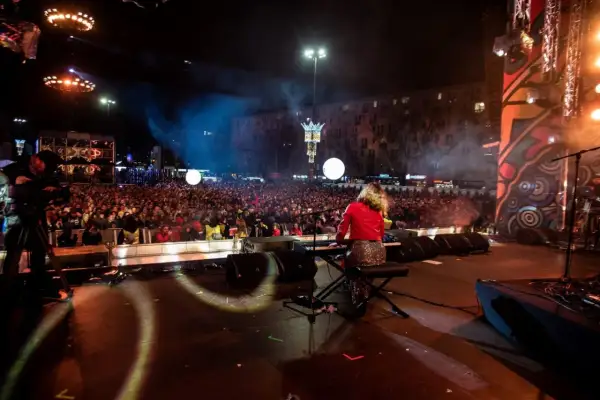 Koncert na scenie z klawiszowcem, tłum ludzi na placu, kamery, światła i dym.
