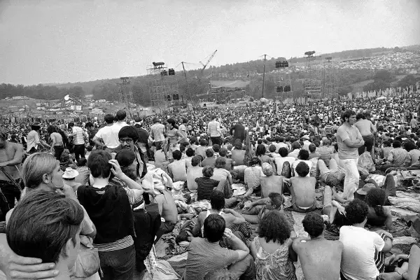 Tysiące ludzi siedzi na trawie, patrząc na scenę festiwalu. To właśnie Woodstock, gdzie jest to legendarne wydarzenie.