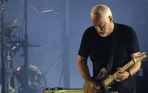 David Gilmour gra na gitarze elektrycznej na tle sceny z dymem i sprzętem muzycznym.