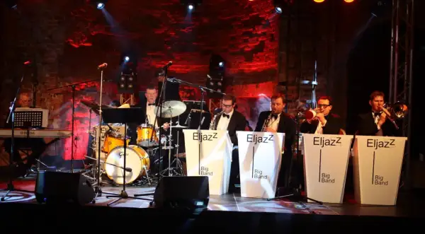 Zespół jazzowy gra na scenie, przygotowując się do sylwestrowych koncertów. Muzycy w eleganckich strojach, instrumenty i czerwone światła tworzą wyjątkową atmosferę.