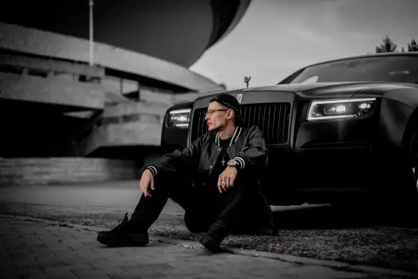 Mężczyzna w czapce i kurtce siedzi obok czarnego Rolls-Royce'a na tle betonowej budowli.