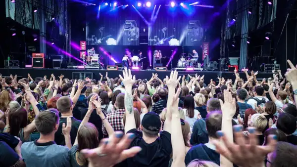 Koncert na żywo, tłum ludzi z podniesionymi rękami, scena z zespołem w tle, fioletowe światła.