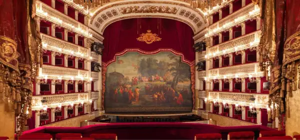 Wnętrze luksusowego teatru operowego z czerwonymi lożami, złotymi zdobieniami i malowidłem na scenie.