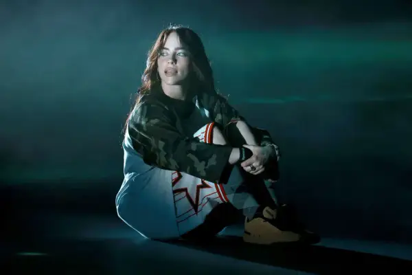 Billie Eilish siedzi na scenie w stroju sportowym, wpatrując się w bok.