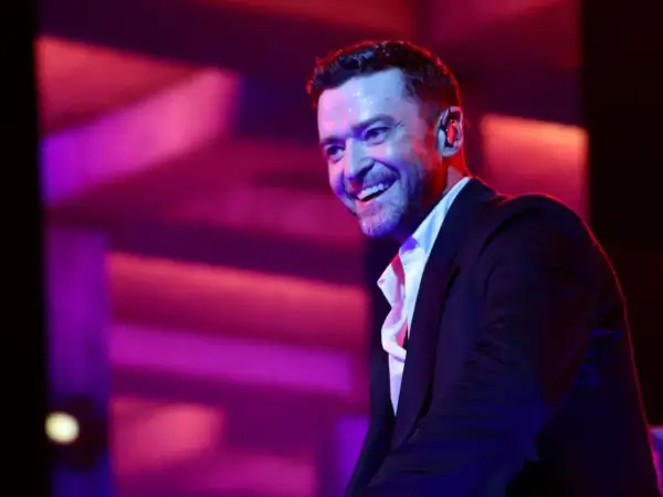 Justin Timberlake w czarnym garniturze i białej koszuli, uśmiecha się na tle fioletowo-różowego oświetlenia scenicznego.
