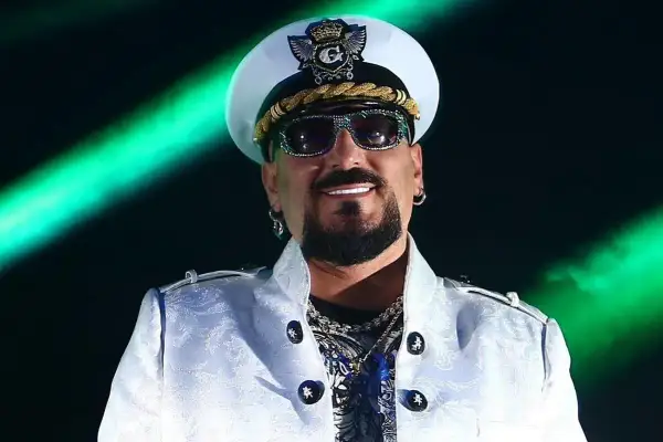 Gigi D'Agostino w białej marynarce i czapce, w okularach przeciwsłonecznych, na tle zielonych świateł.