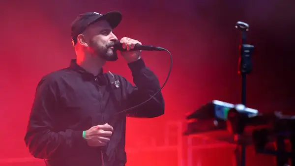 Wokalista z brodą w czapce i kurtce śpiewa do mikrofonu na tle czerwonych świateł. To część tauron nowa muzyka line-up.