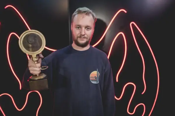 Mężczyzna w granatowej bluzie trzyma złotą statuetkę na tle czerwonych neonów.