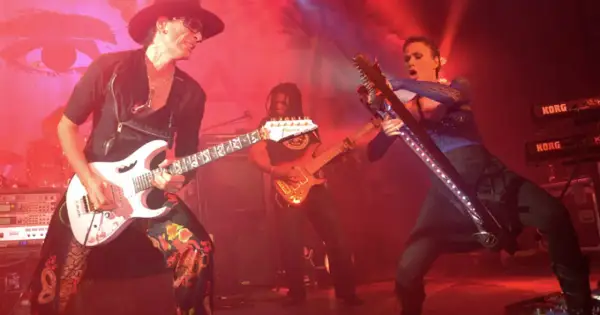 Trzech gitarzystów na scenie, jeden w kapeluszu gra na białej gitarze, drugi w tle na pomarańczowej, trzeci na futurystycznej.