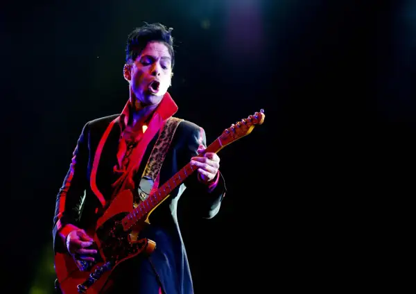 Prince, jako prince opener, gra na gitarze w czerwonej koszuli i czarnej marynarce, oświetlony kolorowymi światłami sceny.