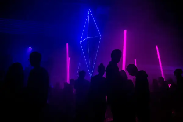 Sylwetki ludzi na tle niebiesko-fioletowych świateł i geometrycznej instalacji świetlnej.