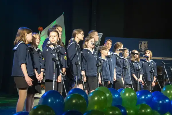 Dzieci w mundurkach śpiewają na scenie, przed nimi mikrofony i balony.