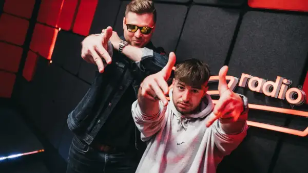 Dwóch mężczyzn pozuje na tle studia radiowego z logo "Zet Radio", wskazując palcami w stronę widza.