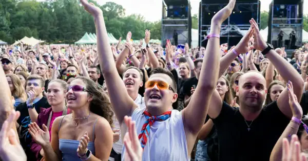 Tłum na koncercie cieszy się letnie brzmienia. Ludzie klaszczą, podnoszą ręce, a niektórzy mają kolorowe okulary i chusty.