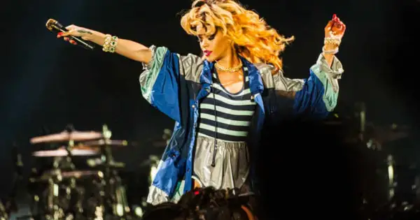 Rihanna na koncercie w Polsce, w bluzie z kolorowymi pasami, śpiewa z mikrofonem w dłoni.