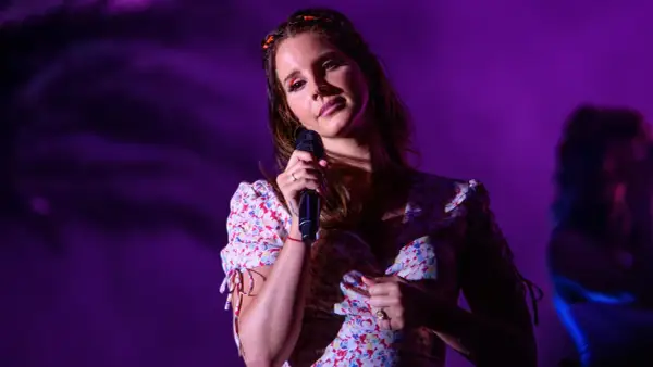 Lana Del Rey śpiewa na scenie w kwiecistej sukience, oświetlona fioletowym światłem.
