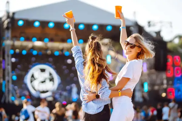 Dwie kobiety z podniesionymi rękami i kubkami na tle sceny festiwalowej.