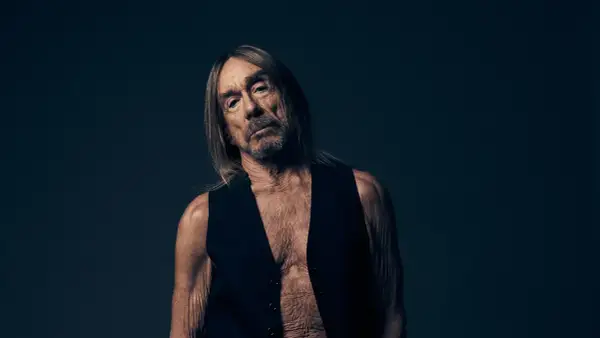 Iggy Pop w czarnej kamizelce, z długimi włosami i zmęczoną miną, na ciemnym tle.
